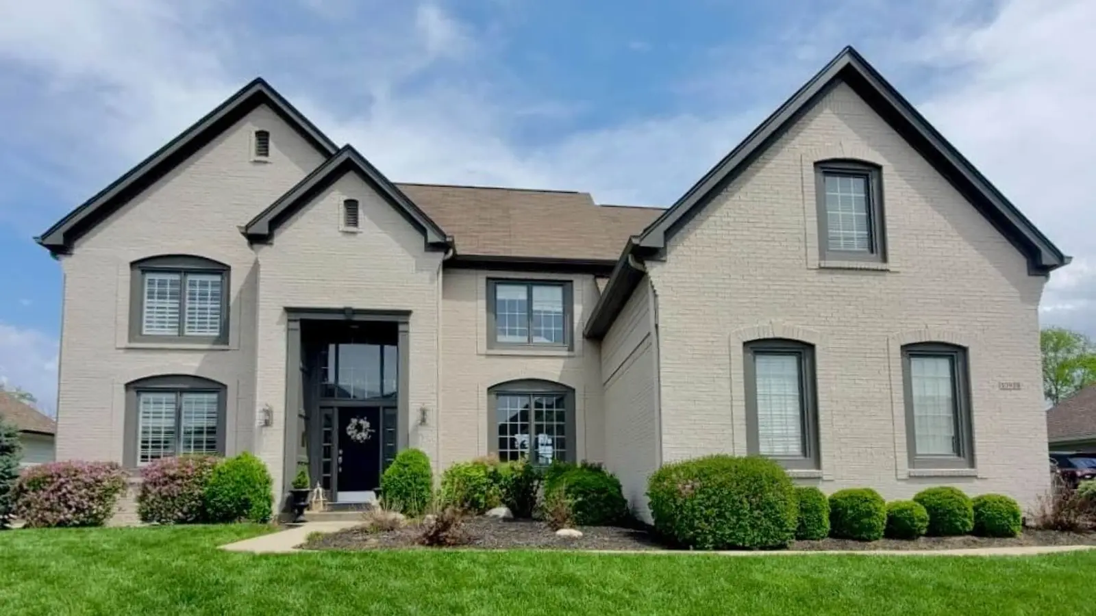 A large, two-story residential home showcasing a professional Indianapolis exterior painting project by Guy Painting. The exterior features painted light gray brick walls that provide a clean, modern foundation. The Guy Painting team applied a high-contrast dark gray finish to the prominent window frames, gutters, and trim along the roofline. This sophisticated color scheme is anchored by a bold black front door with a decorative wreath and matching dark portico, all framed by manicured green shrubs and a lush front lawn.