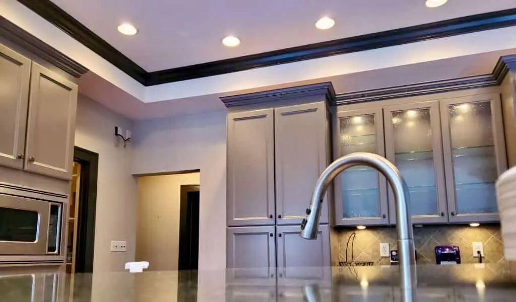 A detailed low-angle view of a Geist kitchen renovation, highlighting the expert finish achieved during a painting interior of kitchen cabinets project. The image focuses on the upper section of a professional cabinetry installation, featuring tall pantry units and glass-front cabinets refinished in Sherwin-Williams Gauntlet Gray. The guy painting team applied a flawless, high-durability coating that extends to the crown molding and matches the recessed ceiling tray's dark trim. A modern brushed nickel gooseneck faucet and integrated stainless steel microwave are visible in the foreground, complementing the sophisticated gray tones and precision craftsmanship provided by the guy painting crew.