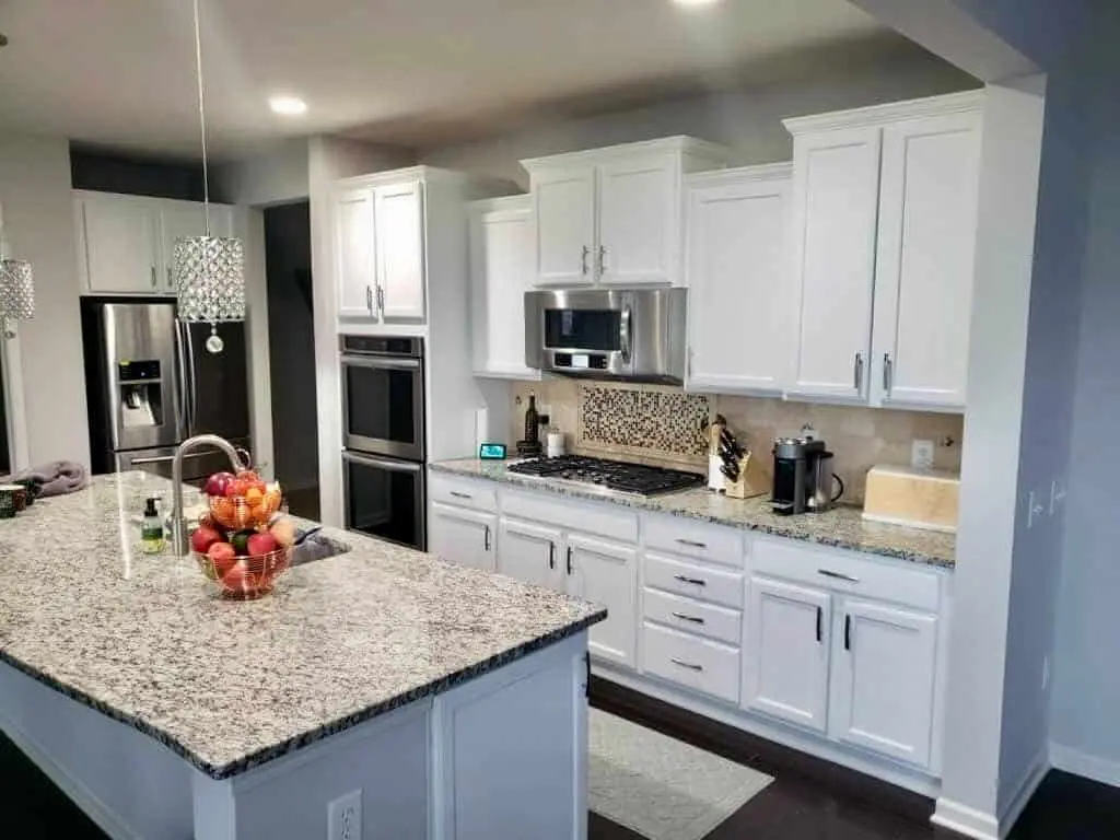 A brightly lit, modern kitchen in Geist showing the completed results of painting kitchen cabinets white after a professional refinishing by the guy painting team. The image features a large central island with a granite countertop and a bowl of fresh fruit, leading to the main cabinetry wall. The shaker-style upper and lower cabinets are finished in a clean, durable white that coordinates perfectly with the stainless steel double ovens, microwave, and refrigerator. A detailed mosaic tile backsplash behind the gas cooktop and elegant crystal pendant lighting complete the sophisticated look of this kitchen transformation by guy painting.