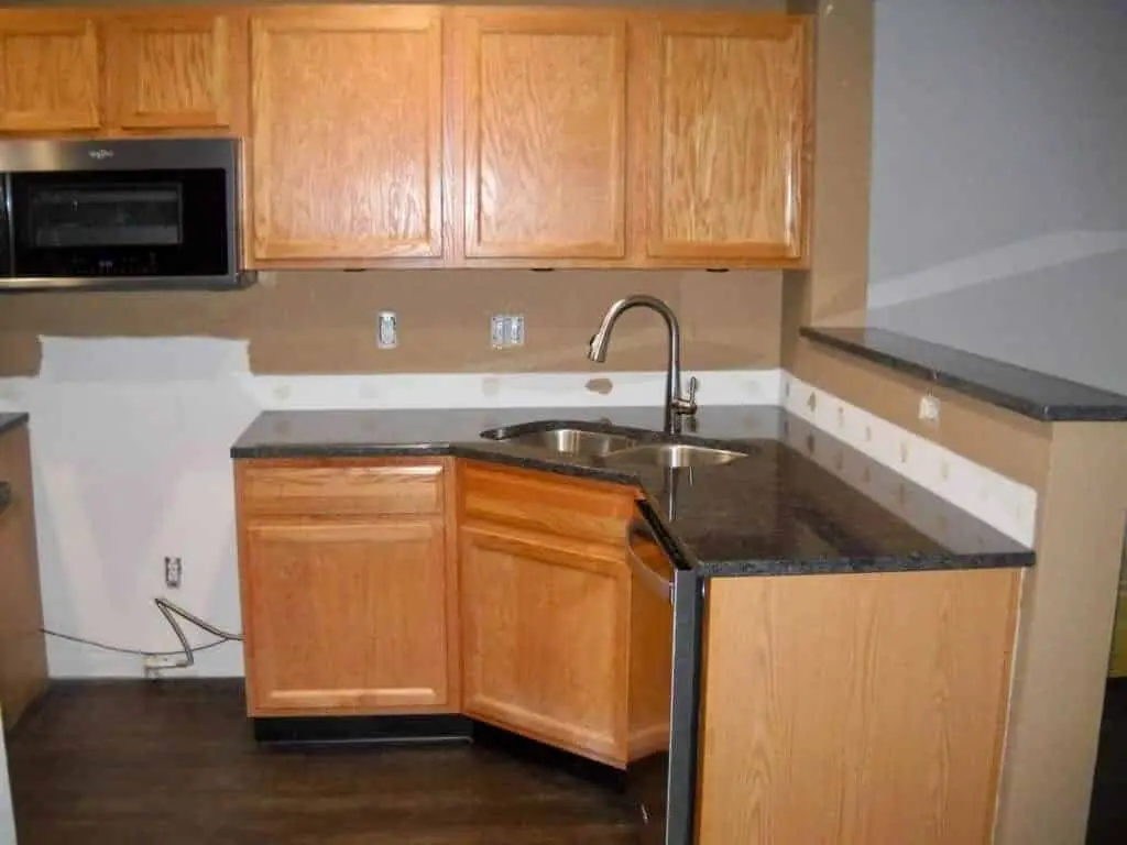 A kitchen preparation scene in a Geist home, showing the original honey oak cabinetry before a professional painting inside kitchen cabinets service. The image features a corner sink area with dark granite countertops and a stainless steel undermount sink. The traditional golden oak upper and lower cabinets are shown with hardware removed, illustrating the detailed surface preparation and masking required by the guy painting team. White filler and sanding marks are visible on the beige backsplash area, documenting the thorough repair and prime phase of this high-quality kitchen cabinet transformation.