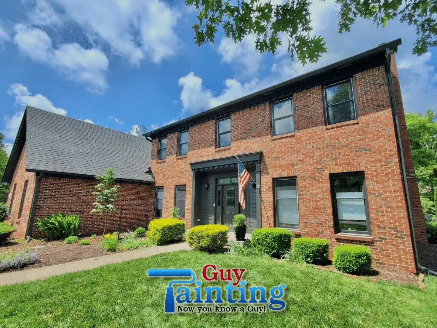 A front-view photograph of a large two-story red and brown brick residential home featuring a professional Indianapolis exterior painting update by Guy Painting. The image highlights a modern color scheme where all window frames, gutters, and the prominent front entryway are finished in a deep black or dark charcoal. An American flag is proudly displayed near the dark-painted front door, which is framed by a sleek, updated portico. The vibrant green lawn and manicured shrubs in the foreground emphasize the enhanced curb appeal provided by the Guy Painting team's meticulous trim work.