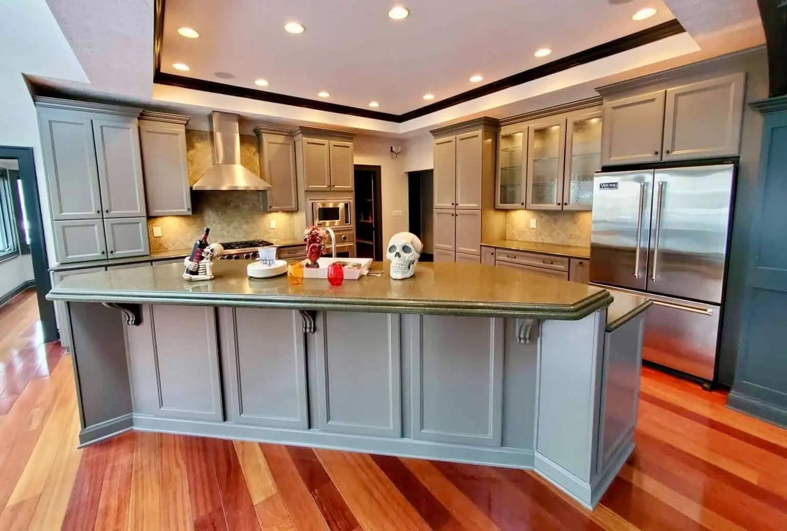 A polished "After" view of a high-end kitchen transformation, showcasing the expert results of painting old kitchen cabinets by the Guy Painting crew. The cabinetry, including the large, multi-angled kitchen island and full-height pantry units, is finished in a smooth, professional coat of Gauntlet Gray. The sophisticated dark gray tone creates a modern, upscale feel that contrasts beautifully with the warm, glossy hardwood floors. This comprehensive refinishing project by Guy Painting features integrated stainless steel appliances, a sleek range hood, and specialized glass-front upper cabinets with interior lighting, all unified by the precision paint application.