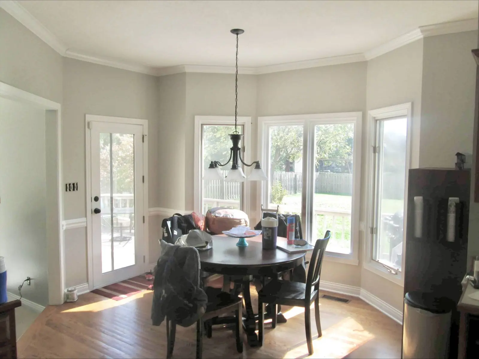 A bright and airy "After" shot showcasing a professional house interior painting Westfield project by Guy Painting. The image highlights a kitchen breakfast nook and dining area where the once dark, stained wood trim has been completely transformed with a crisp, semi-gloss white paint. The white finish extends across the crown molding, window casings, door frames, and chair rails, perfectly complementing the light gray walls and hardwood flooring. This interior update by Guy Painting demonstrates how a professional color change on trim can modernize a home and maximize the natural light in a living space.