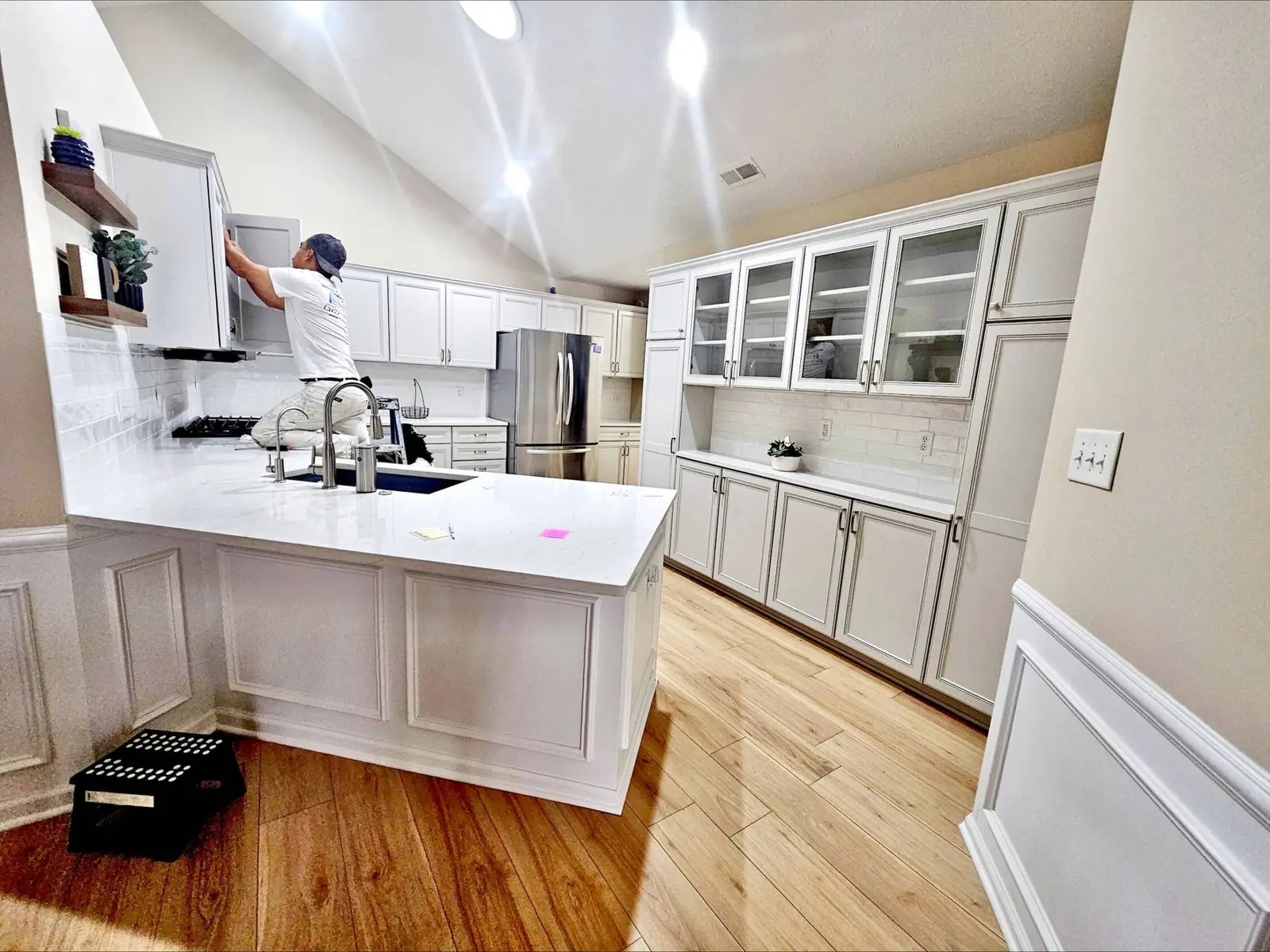 A wide-angle view of a completed white painted oak kitchen cabinets Indianapolis project in Geist. The image shows a professional painter from the Guy Painting team meticulously installing hardware on a freshly refinished upper cabinet. The entire kitchen has been updated from original wood to a bright, durable white finish, featuring a large island, glass-front hutch, and elegant crown molding. The crisp white cabinetry perfectly complements the light wood-look flooring and white subway tile backsplash, showcasing the high-quality craftsmanship and modern aesthetic delivered by the Guy Painting crew.