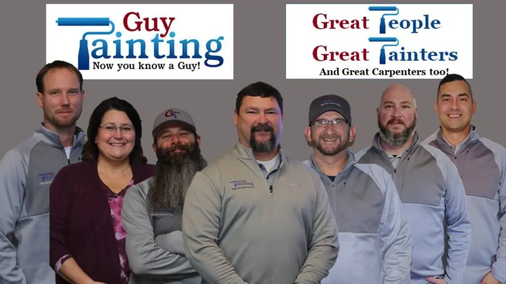 A group portrait of the professional painters indianapolis team from guy painting in geist. The image features seven smiling team members, including several men in grey branded quarter-zip pullovers and caps alongside a woman in a maroon shirt. Above the team, two logos are displayed: the official guy painting logo and a secondary banner that reads "Great People Great Painters And Great Carpenters too!" This photo highlights the friendly, expert crew dedicated to high-quality home transformations and reliable service across the indianapolis area.